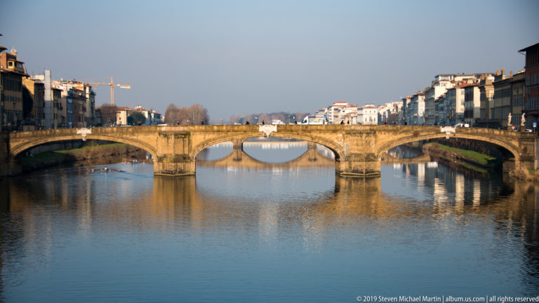 Three-arched Ponte Santa Trinita crossing the calm Arno River between rows of historic riverside buildings in Florence.