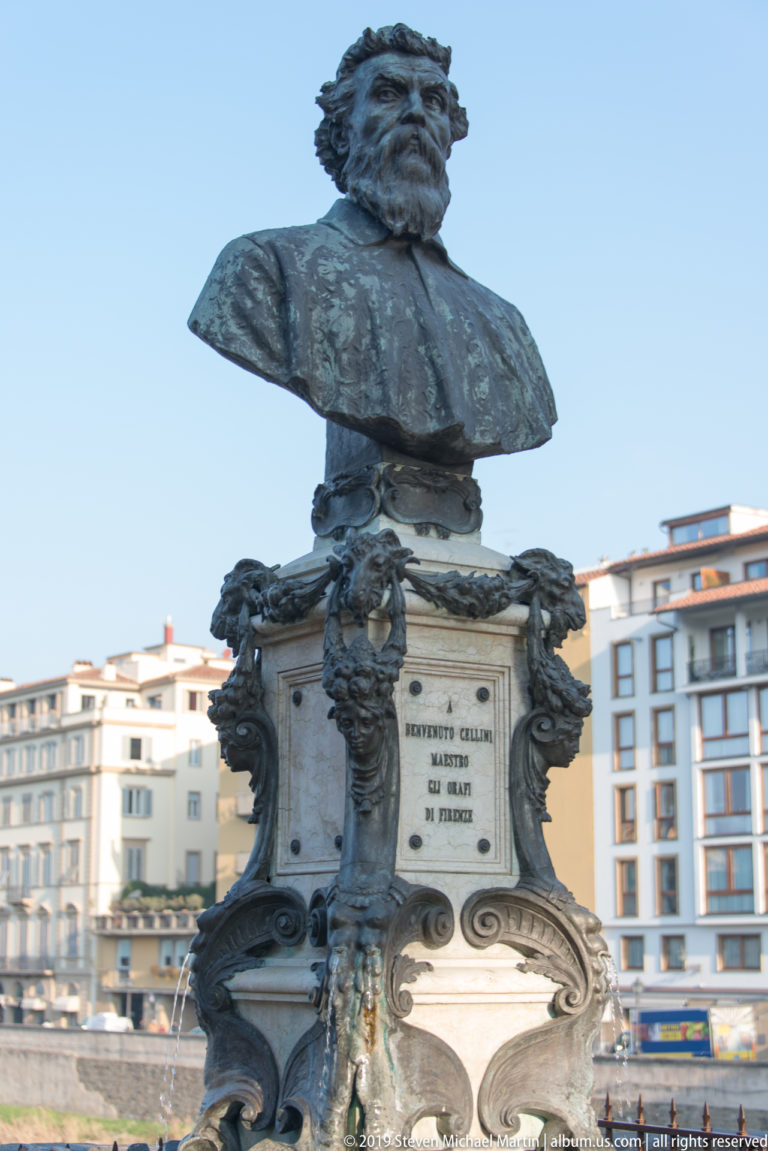 Bronze bust of Benvenuto Cellini above an ornate carved stone pedestal with inscription, masks, and scrollwork against blue sky.