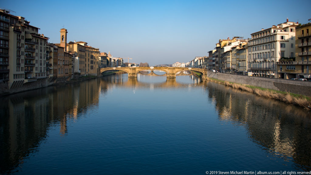 Centered view along the calm Arno River toward three-arched Ponte Santa Trinita, with historic palazzi lining both banks.