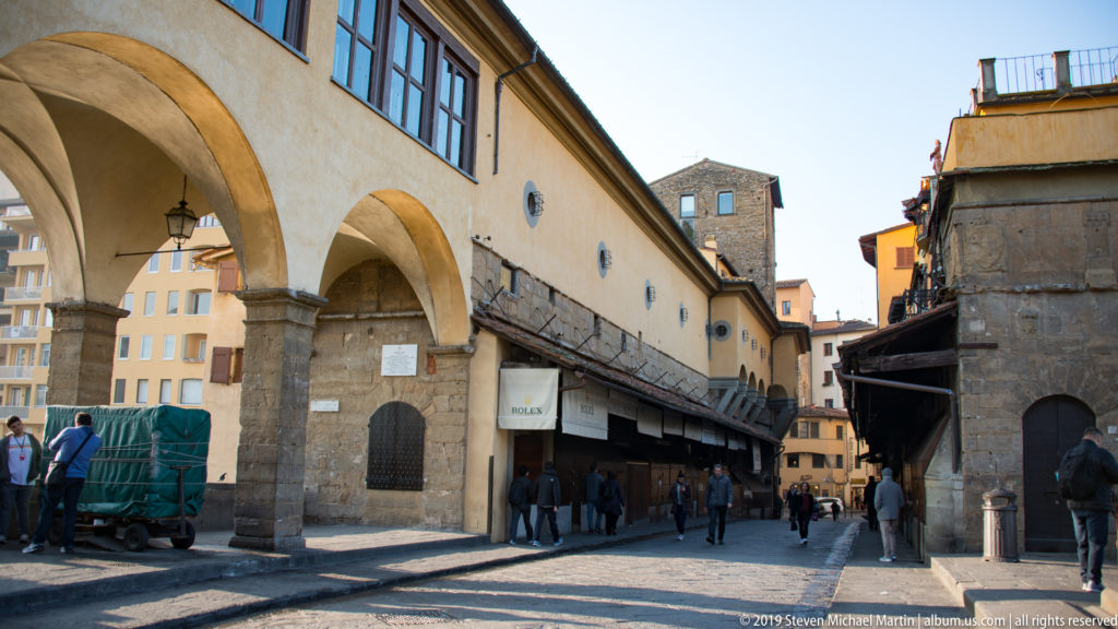 Arcaded ochre building with shops, pedestrians, stone piers, and a masonry tower near the approach to Ponte Vecchio.