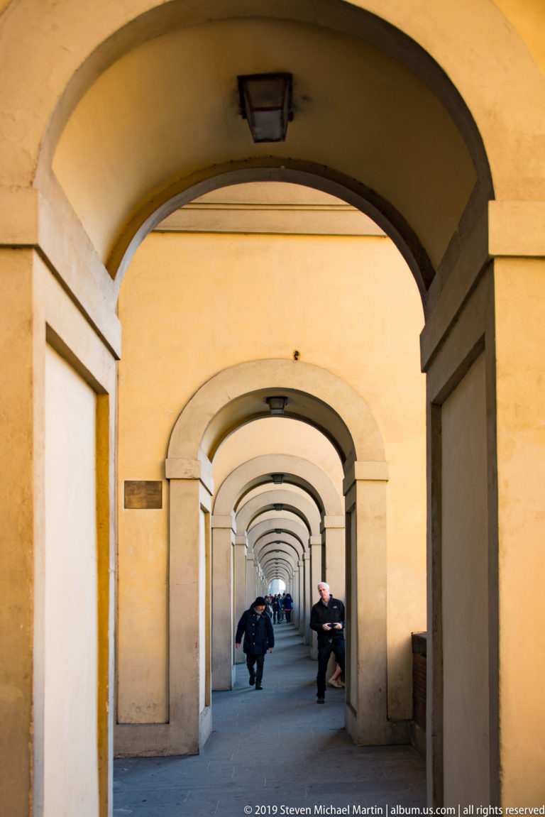 People sitting and walking beneath a long sequence of pale rounded arches in a covered Florentine passage.