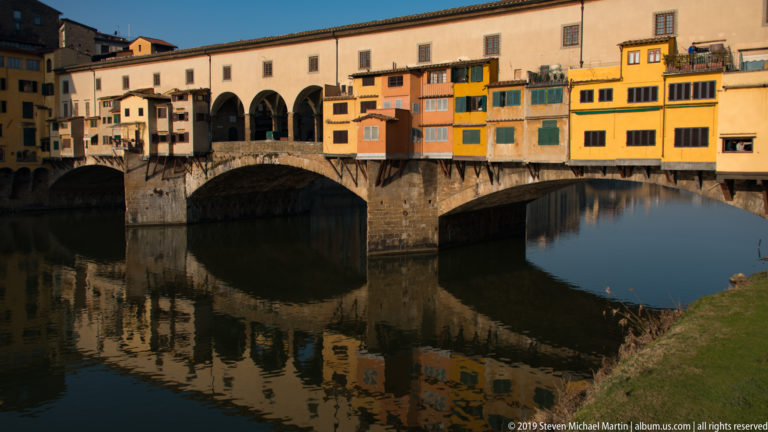 Ponte Vecchio with colorful projecting shops and broad stone arches reflected in the dark, calm Arno River.