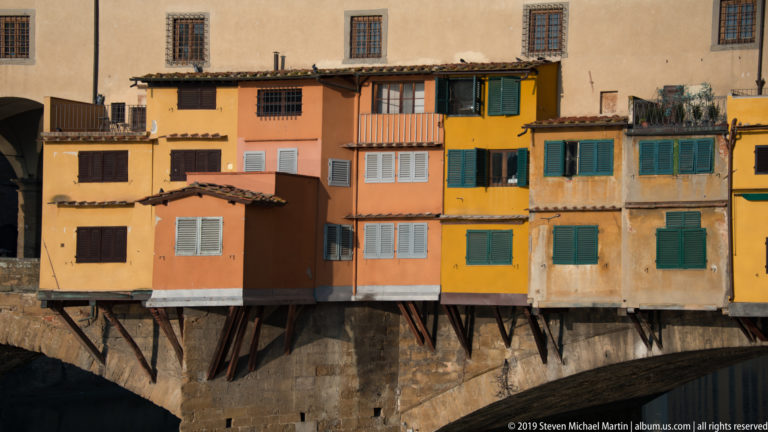 Colorful projecting shop rooms with shutters and angled supports built along the side of Ponte Vecchio above a stone arch.