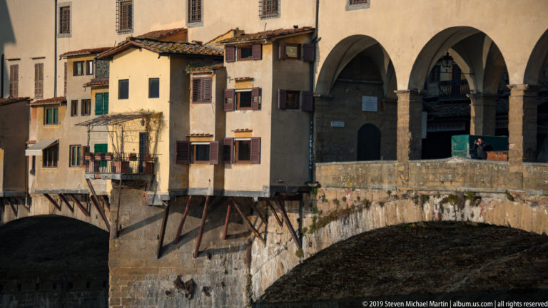 Oblique view of Ponte Vecchio with projecting ochre shop rooms, stone arches, supports, and an open riverside arcade.