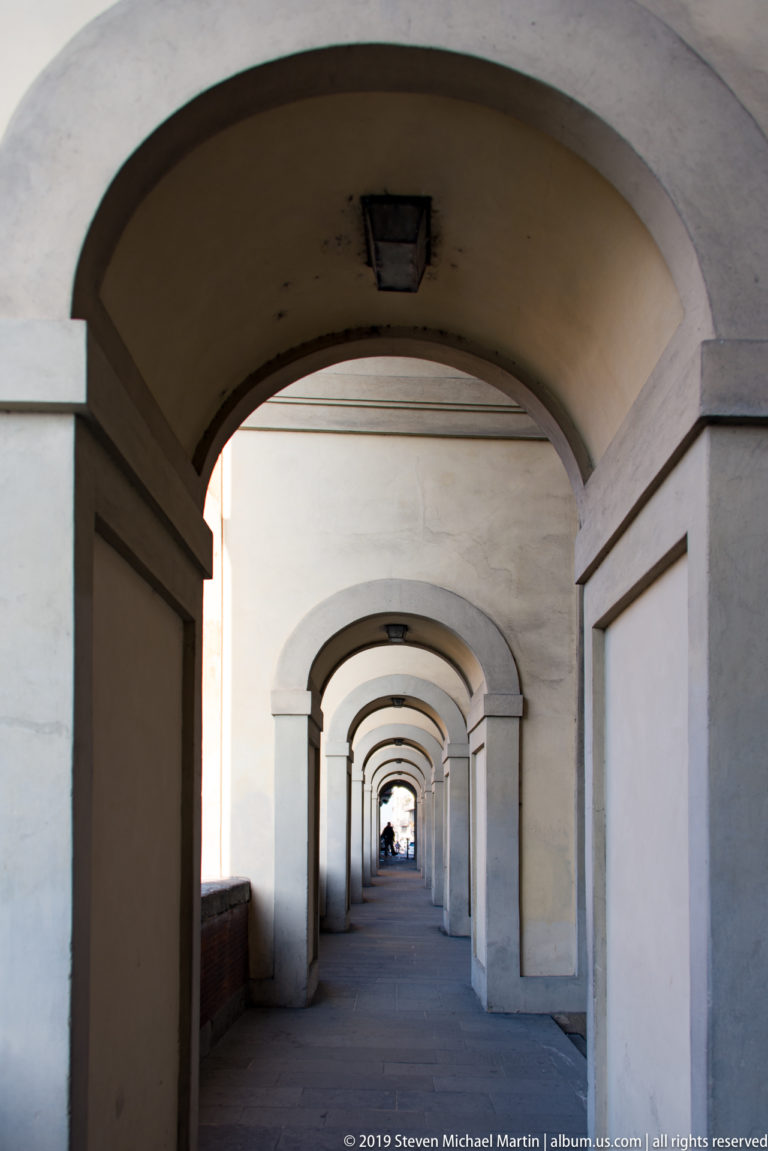 Long pedestrian arcade of repeated pale rounded arches receding toward distant figures in Florence.