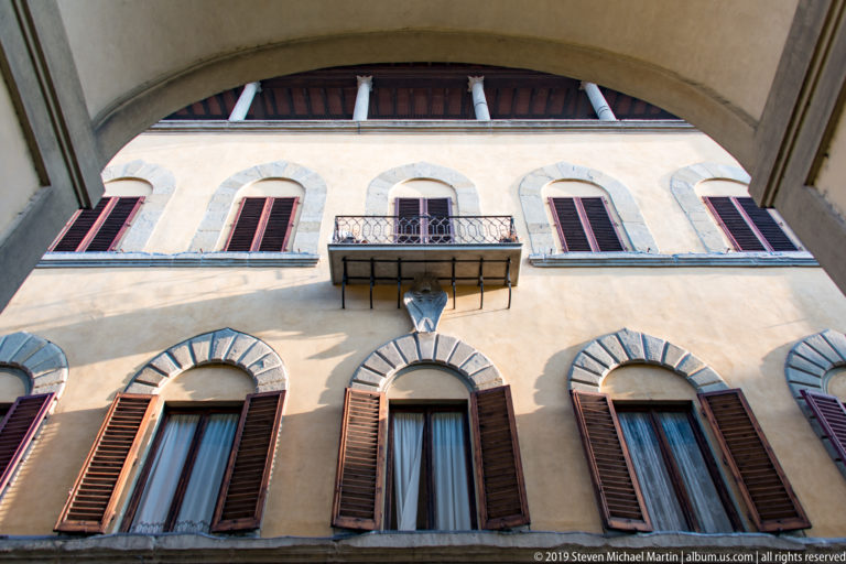 Upward view of ochre Florentine façade with gray stone window arches, dark shutters, and a small wrought-iron balcony.
