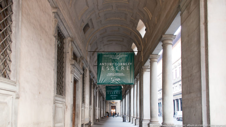 Long Uffizi colonnade with repeated stone columns, coffered vaulted ceiling, and green Antony Gormley exhibition banners.