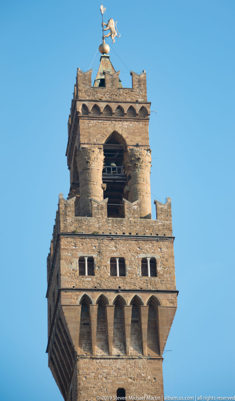 Upper stages of a medieval stone tower with arched openings, crenellations, a large bell, and metal weather vane against blue sky.