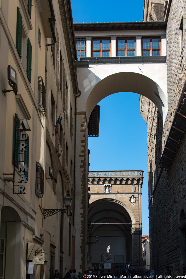 White enclosed passage bridging a narrow Florence street above shops and pedestrians, with another large arch visible beyond.
