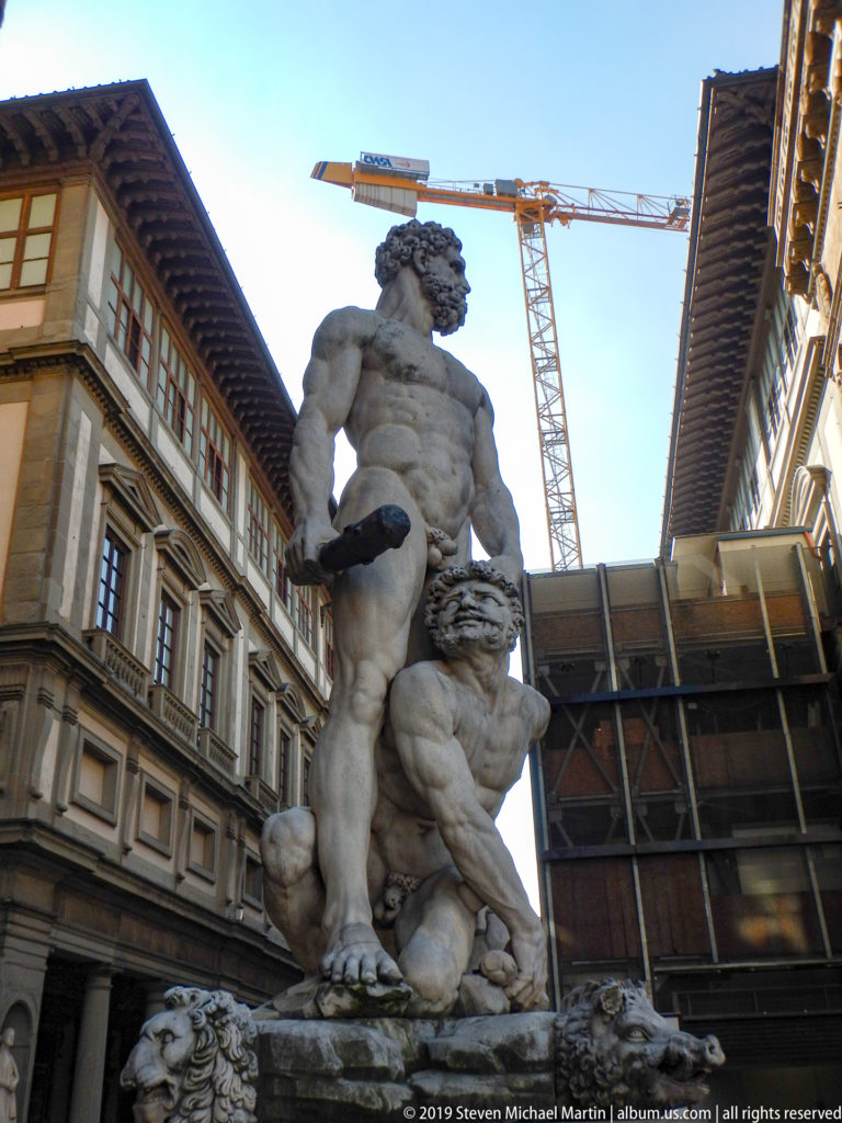 Full marble Hercules and Cacus group seen from below with historic façades, scaffolding, and a construction crane behind.