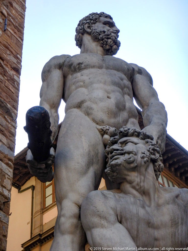 Low-angle close view of marble Hercules holding a club above the lower figure of Cacus against sky and historic façades.