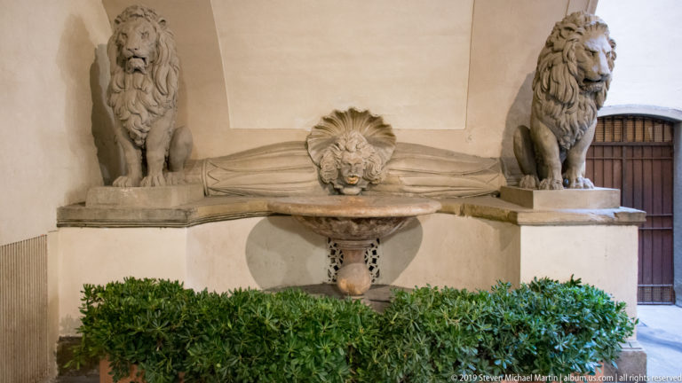 Stone wall fountain with a central lion mask and shallow basin flanked by two seated lion sculptures in an arched recess.