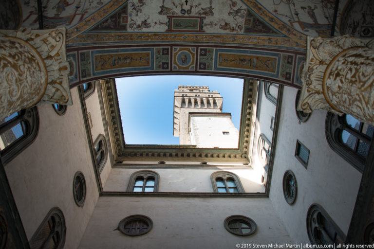 Centered upward view from Palazzo Vecchio’s decorated courtyard toward the Torre di Arnolfo against blue sky.