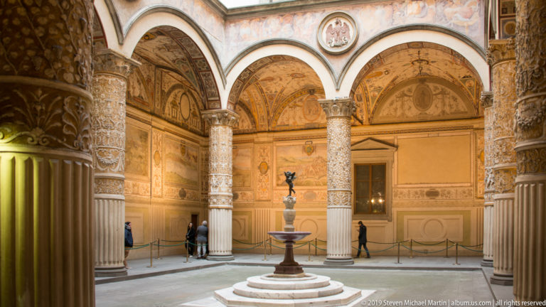Palazzo Vecchio courtyard with arcades, richly ornamented columns and walls, visitors, and a central fountain sculpture.