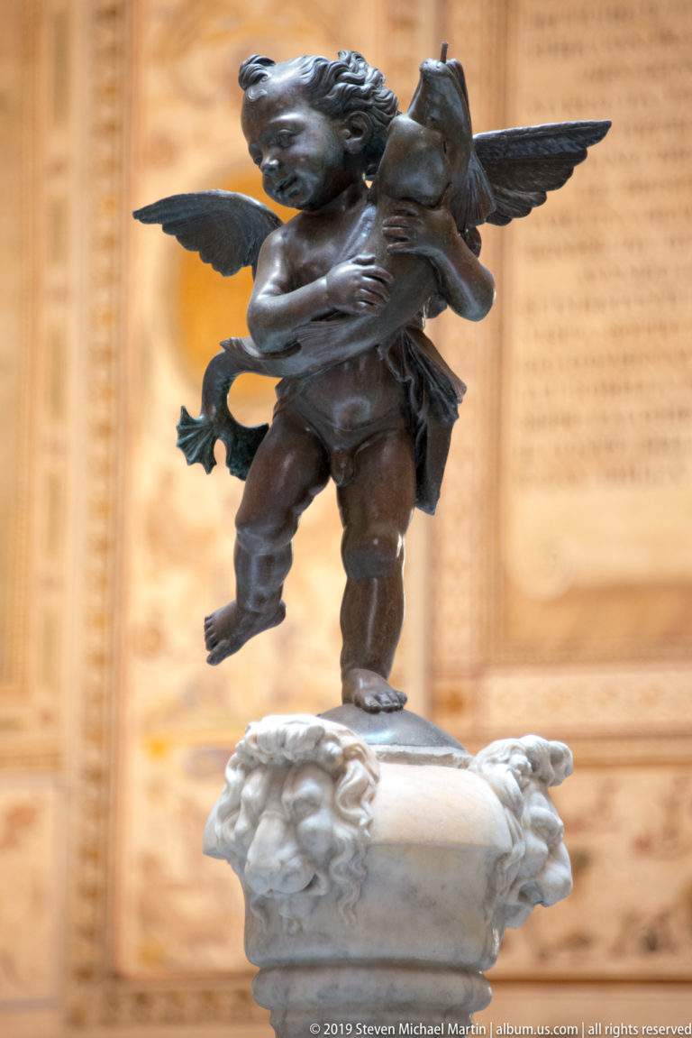 Winged bronze Putto with Dolphin atop a pale fountain basin, with frescoes and an ornate courtyard column behind.
