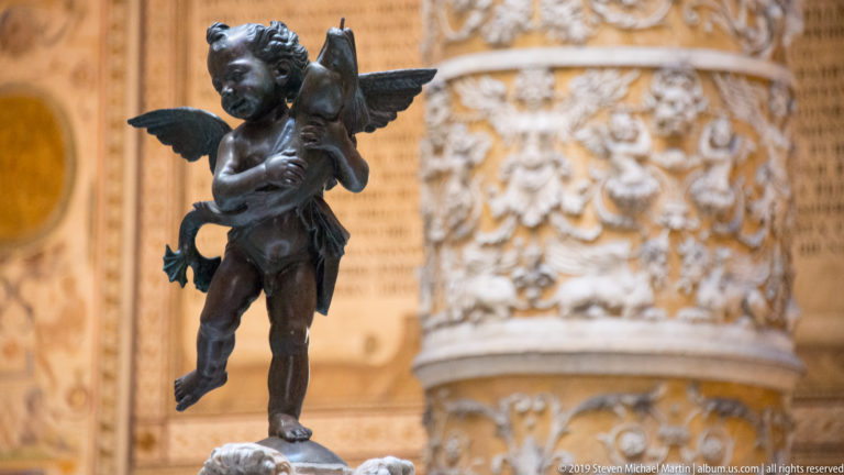 Close frontal view of a winged bronze putto embracing a dolphin, with ornate courtyard decoration blurred behind.