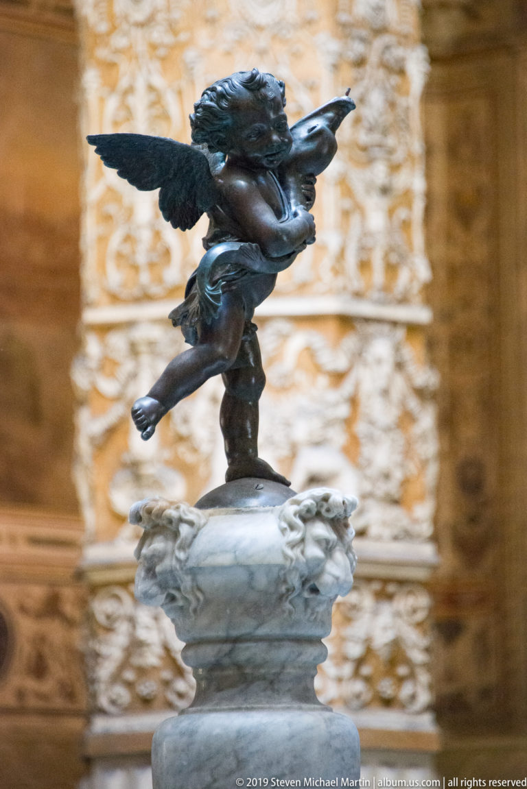 Winged bronze putto twisting around a dolphin atop a pale fountain pedestal in an ornate Florentine courtyard.