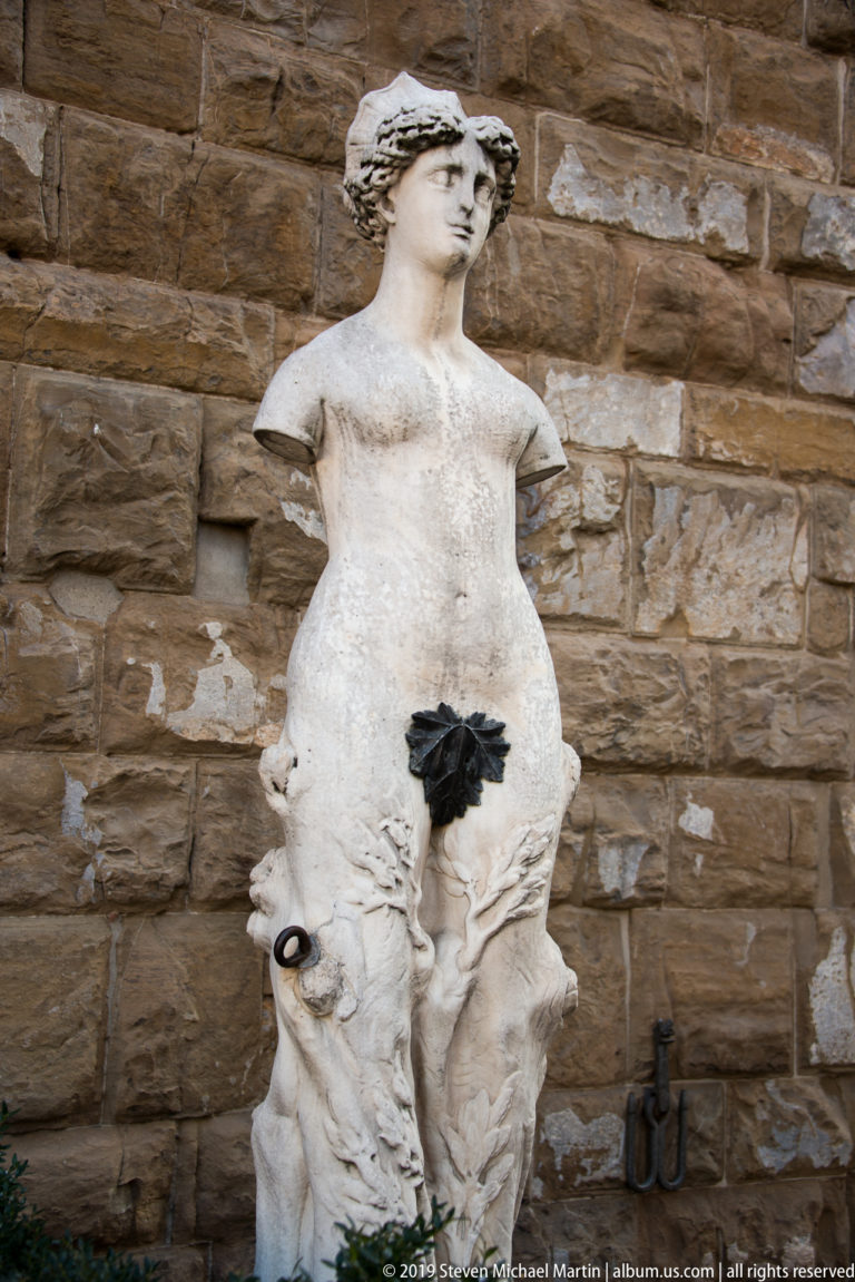 Weathered pale stone female statue with missing arm and drapery around the hips against a rusticated masonry wall.