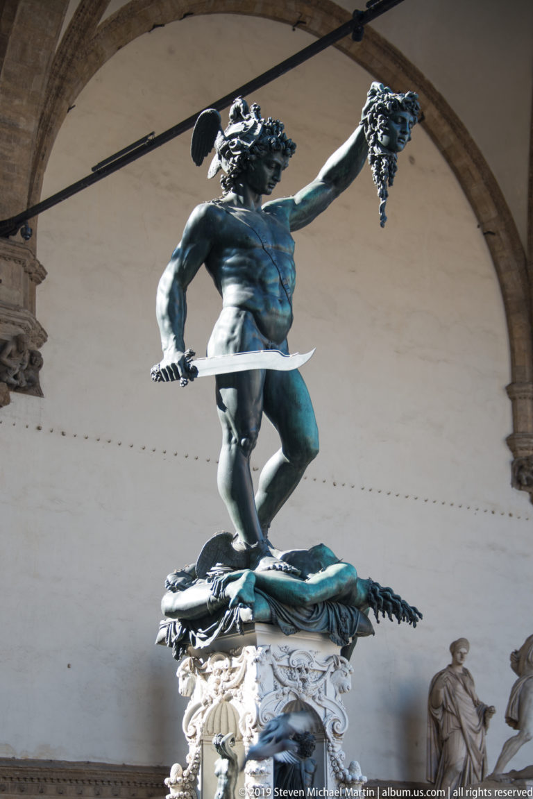 Full bronze Perseus raising Medusa’s head and holding a sword while standing above her body on an ornate pedestal.