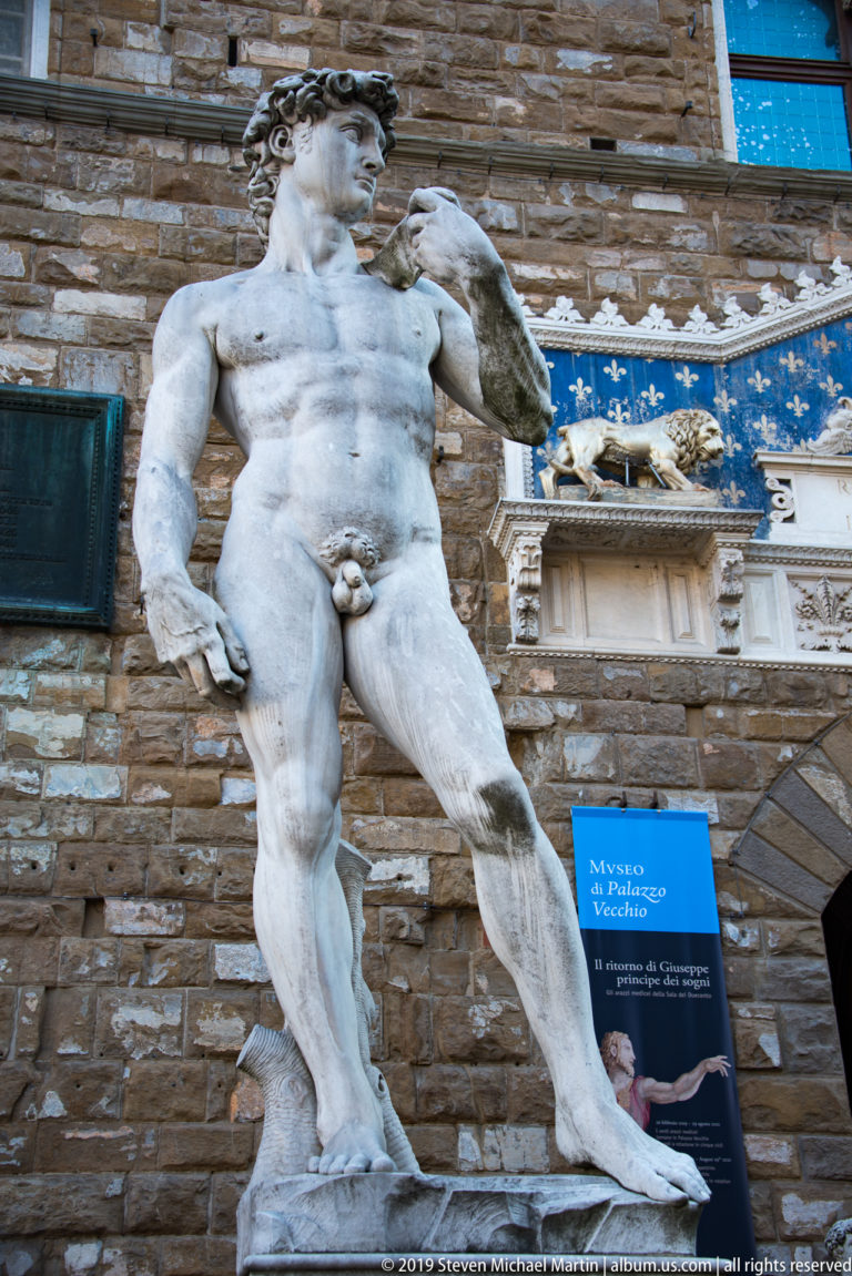 Low-angle full view of the David replica standing before Palazzo Vecchio and its blue heraldic entrance panel.
