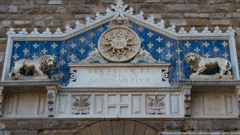 Blue-and-gold heraldic panel with lions, fleurs-de-lis, Christogram, and Latin inscription above Palazzo Vecchio’s entrance.