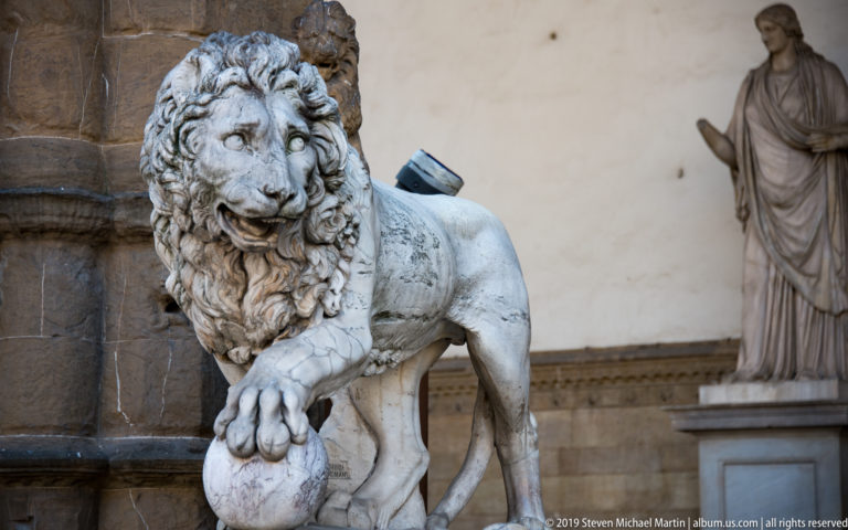 Carved stone lion with a thick mane resting one paw on a rounded shield, with another statue in the background.