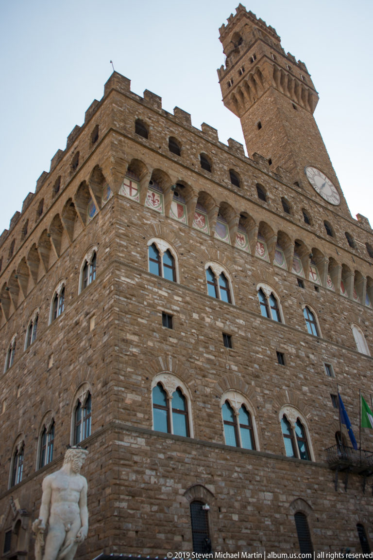 Low-angle view of Palazzo Vecchio’s rusticated stone walls, Gothic windows, painted shields, crenellations, and tower.