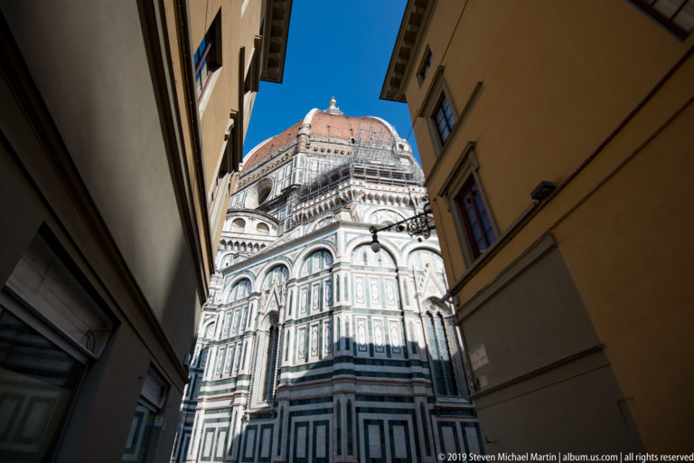 Baptistery of San Giovanni framed by a narrow street, with the terracotta dome of Florence Cathedral rising behind.