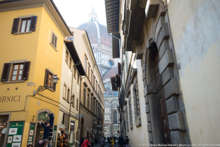 Florence street with pedestrians and ochre façades opening toward the marble cathedral and its large terracotta dome.