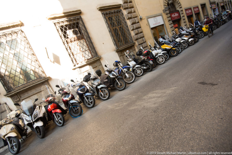 Long diagonal row of parked scooters and motorcycles beside a historic stone building on a Florence street.