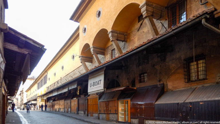 Long diagonal row of closed dark wooden shops beneath ochre arches and heavy stone supports near Ponte Vecchio.