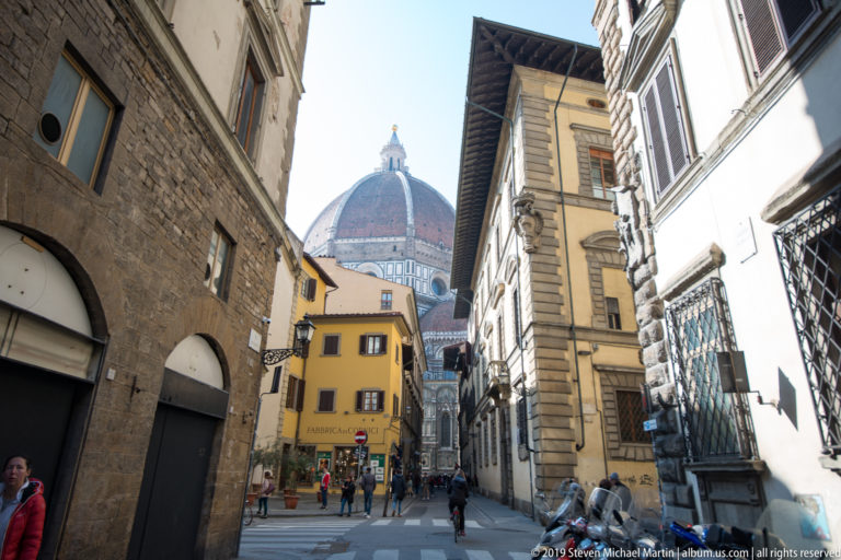 Narrow Florence street leading toward the cathedral, with Brunelleschi’s terracotta dome rising above surrounding buildings.
