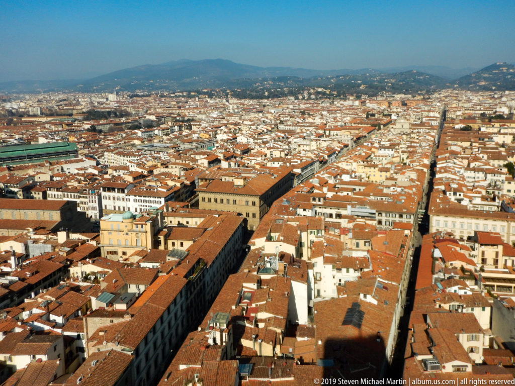 Wide elevated view over Florence’s terracotta rooftops, long straight streets, monumental buildings, and surrounding hills.