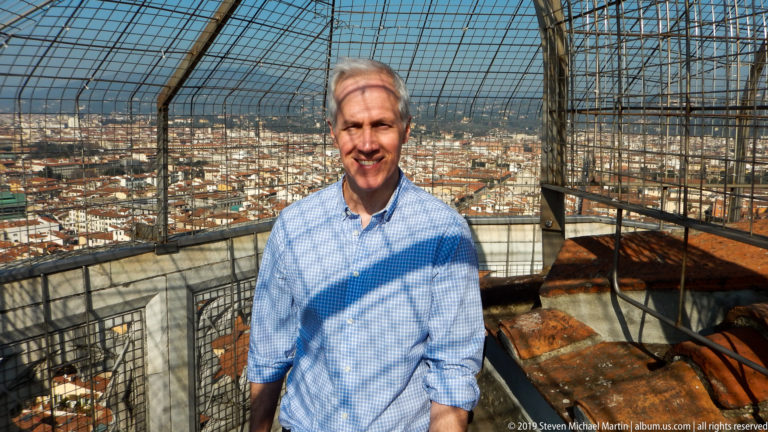 Man standing beside a protective metal enclosure high above Florence, with terracotta rooftops and distant hills behind him.