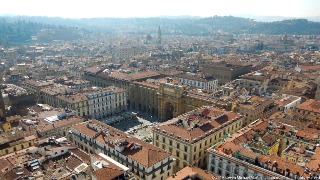 Elevated panorama across central Florence with terracotta roofs, a large rectangular piazza, monumental buildings, and hills beyond.