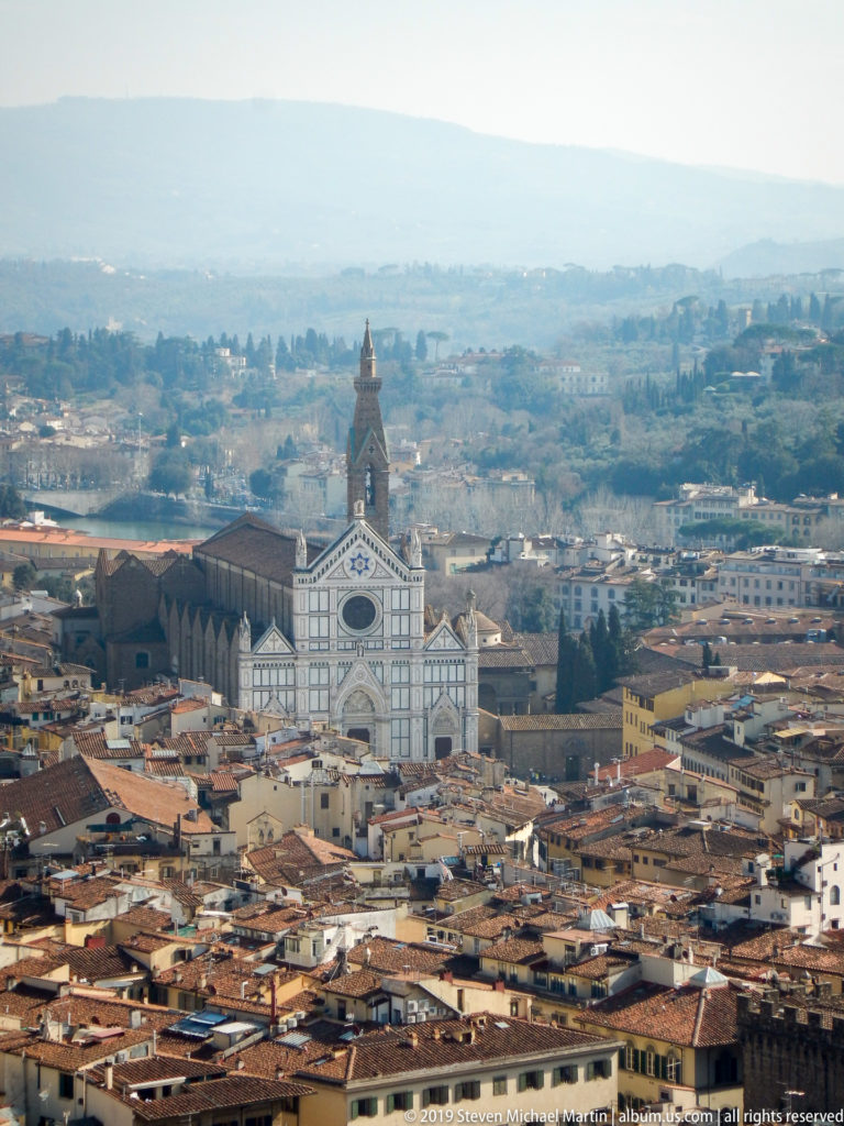 Wide elevated view of Santa Croce with its polychrome façade and campanile surrounded by Florence rooftops.