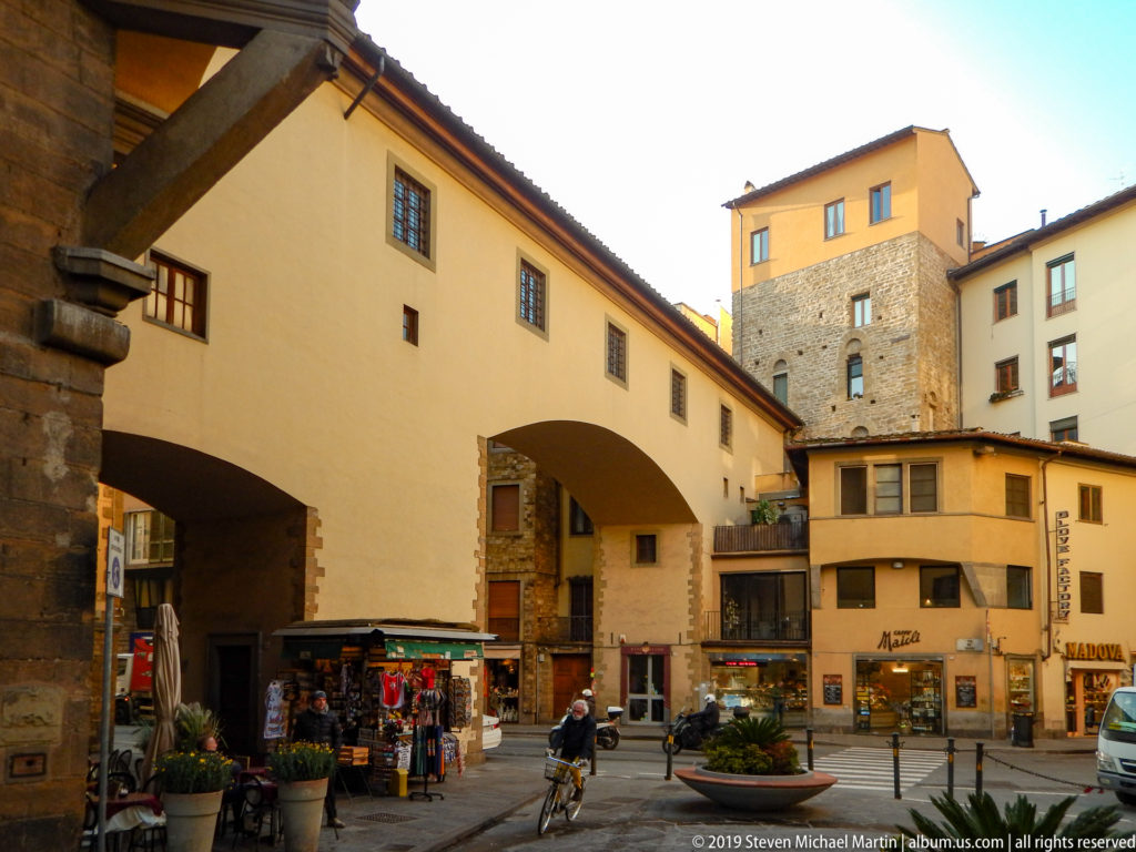 Enclosed passage spanning a small Florence street toward a masonry tower, with shops, bicycles, pedestrians, and ochre buildings.