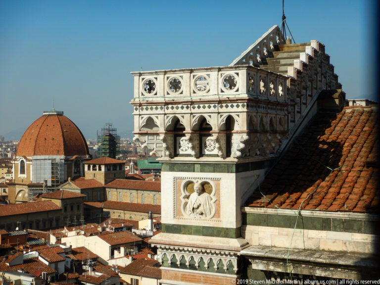 Ornate marble rooftop structure with roundels, arches, and a religious relief, with Florence Cathedral’s dome partly visible at left.