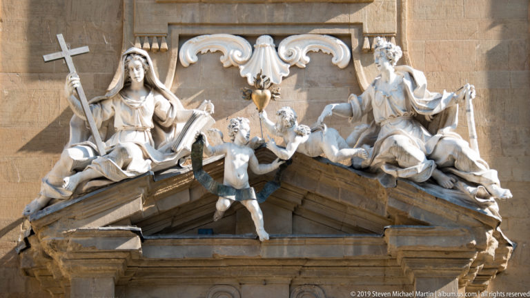 Stone allegorical figures arranged over a broken pediment with scroll ornament on a sunlit Florentine façade.