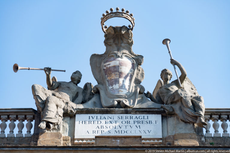 Crowned heraldic shield flanked by seated stone figures with long trumpets above a Latin inscription and balustrade.