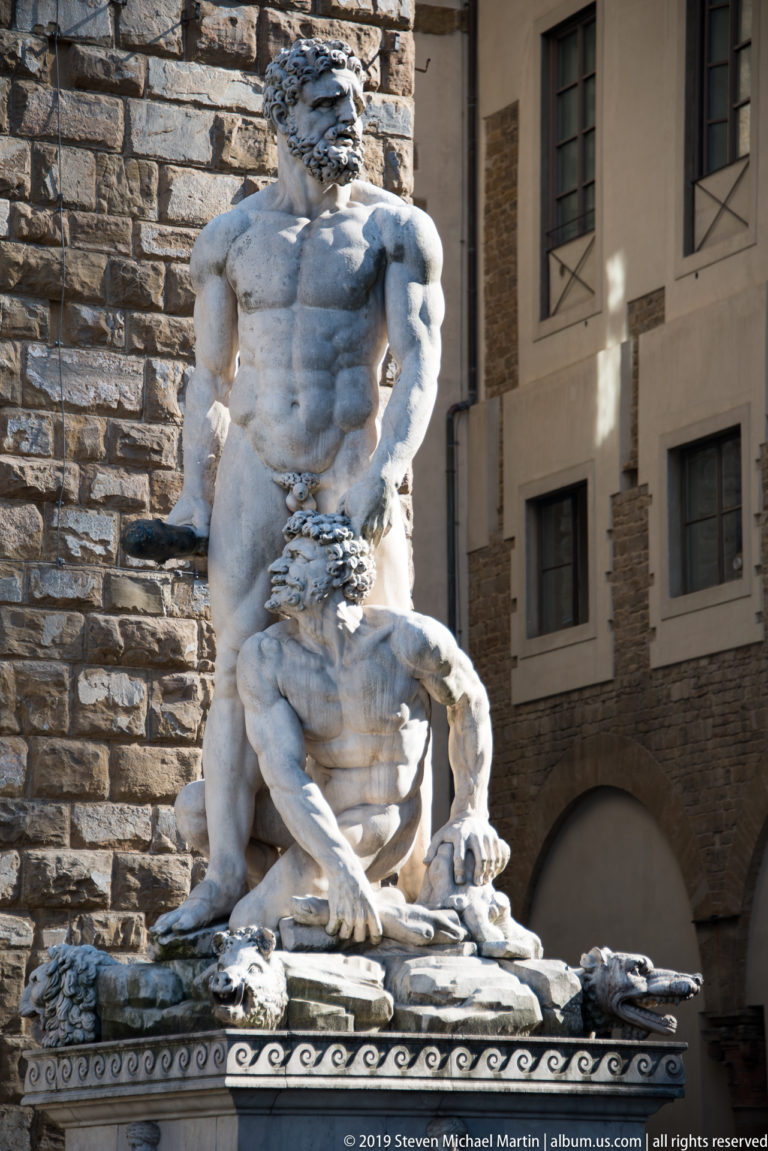 Full marble Hercules and Cacus group in strong sunlight against the rusticated stone wall of Palazzo Vecchio.