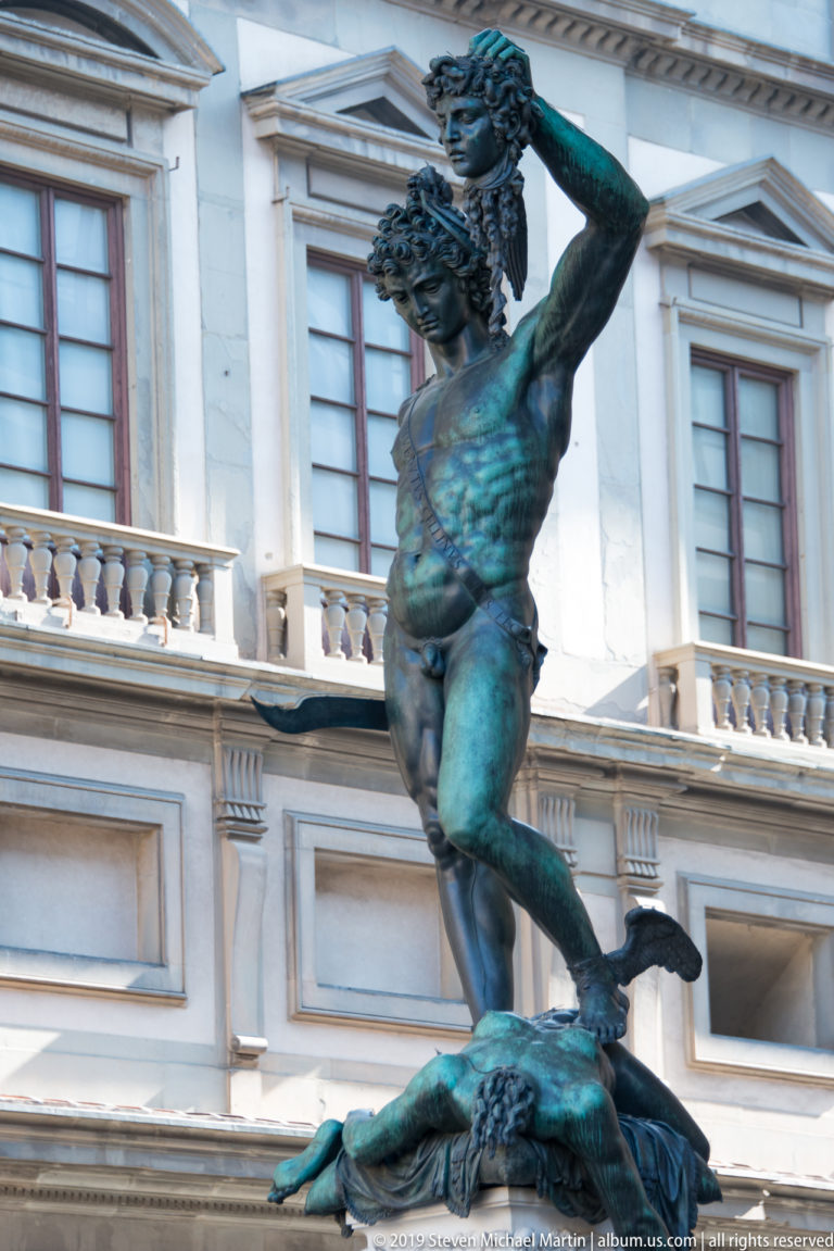 Bronze Perseus raising Medusa’s severed head while standing over her body, with a pale Florentine façade behind.