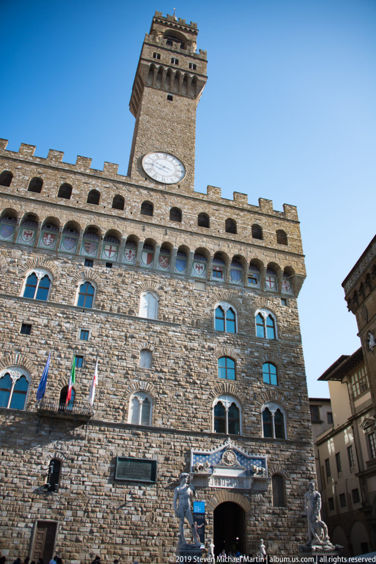Low-angle view of Palazzo Vecchio with rusticated masonry, flags, painted shields, crenellations, and clock tower.