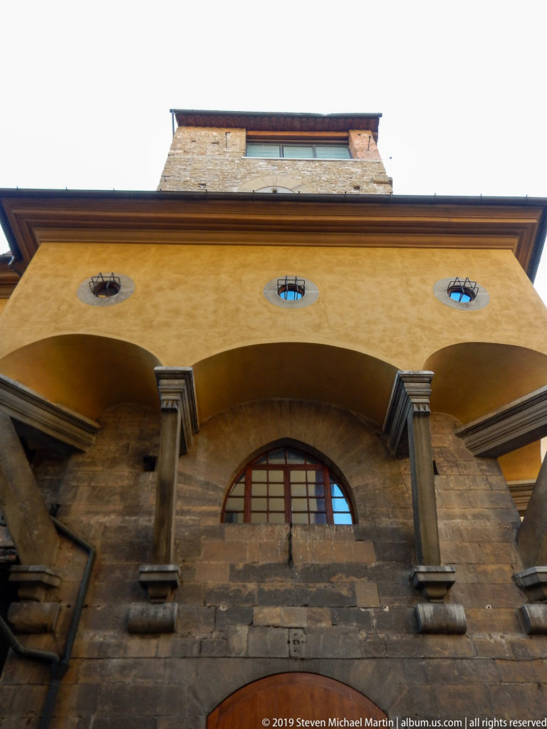 Low-angle view of heavy stone piers supporting ochre arches with circular openings and a rough masonry tower above.