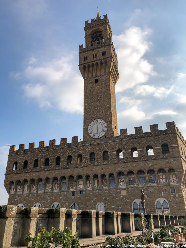 Low-angle view of Palazzo Vecchio’s rusticated stone façade, painted shields, crenellations, clock, and Torre di Arnolfo.