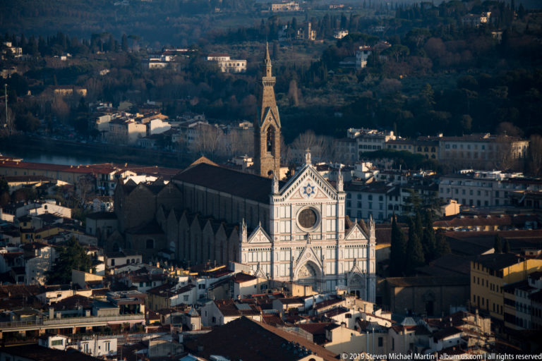 Elevated view of Santa Croce’s polychrome façade, long nave, and pointed bell tower amid Florence’s rooftops.