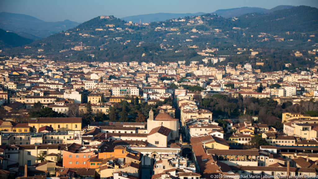 Elevated view of Florence neighborhoods spreading toward wooded hills dotted with villas and houses.
