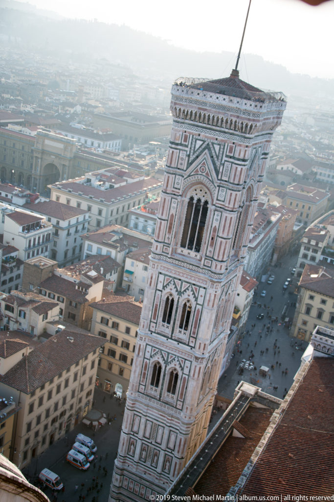 Giotto’s Campanile seen steeply from above, its polychrome marble Gothic stages rising over streets and rooftops.