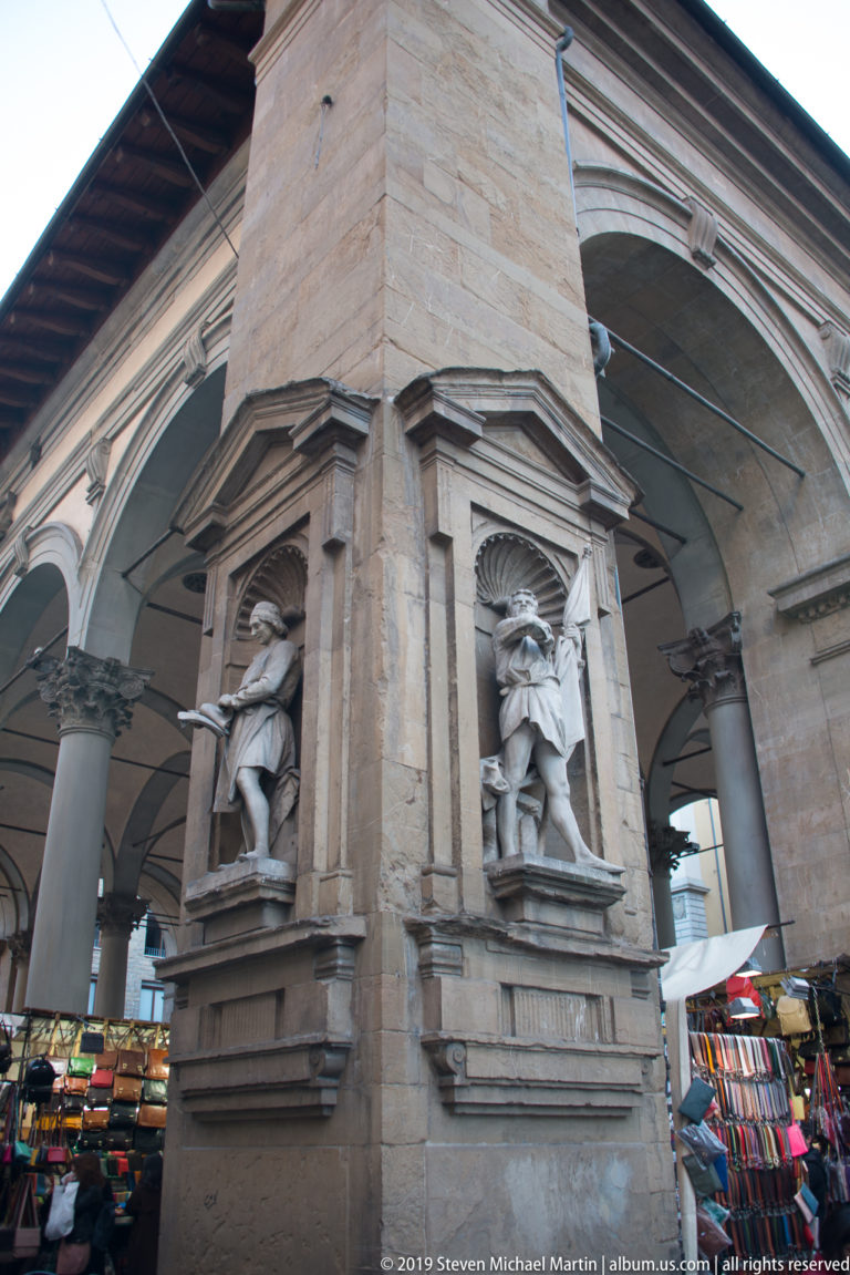 Stone market arcade with two sculpted niche figures on a central pier and colorful merchandise stalls beneath the arches.