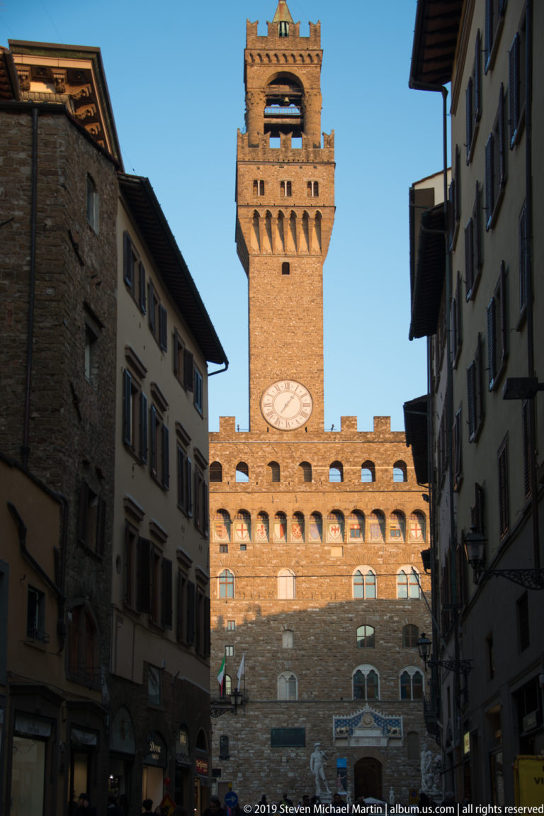 Palazzo Vecchio’s rusticated façade, crenellations, clock, and Torre di Arnolfo seen from a narrow shaded street.